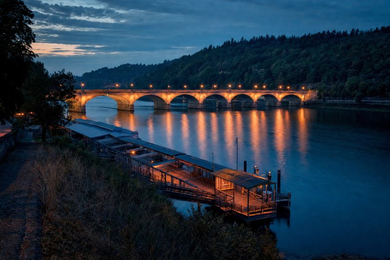 Ein breiter Fluss in der blauen Stunde, eine lange Sandsteinbrücke und Anlegestellen am nahen Ufer, bewaldete Hänge auf der anderen Seite (mit AI erstelltes Bild)