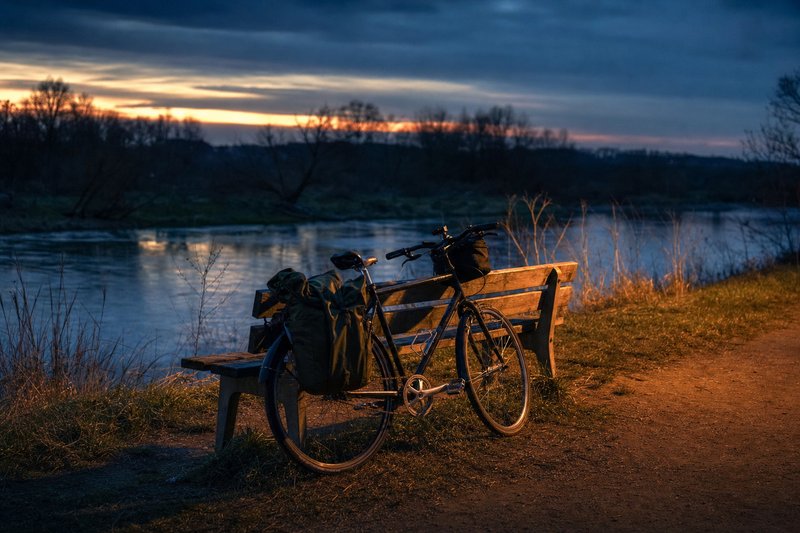 Ein Fahrrad mit Packtaschen lehnt an einer Holzbank auf einer Flusswiese im Abendlicht (mit AI erstelltes Bild)